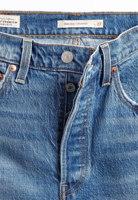 Jean en denim bleu avec braguette à boutons partiellement ouverte, coutures visibles, passants de ceinture, et étiquette de taille indiquant Ribcage Straight 27.
