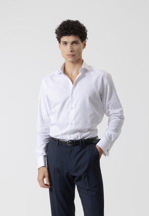 Hombre con cabello rizado, vistiendo una camisa blanca de vestir con botones y pantalones oscuros, de pie con una mano en el bolsillo frente a un fondo liso.