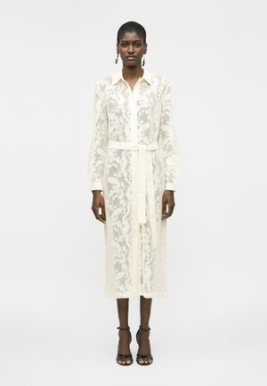 Tumesnaha naine, kes kannab pikka valget pitskleiti krae, vöö ja mustade avatud varvastega kontsakingadega, seisab ühevärvilise valge tausta ees.