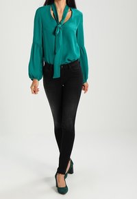 Blusa verde azulada con un lazo en el escote, mangas largas abullonadas, combinada con unos pantalones ajustados negros y zapatos de tacón verdes. Fondo limpio.