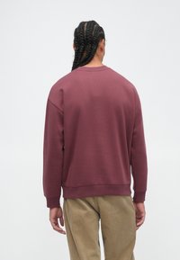 Sweatshirt crewneck bordeaux avec des poignets et un ourlet côtelés, fabriqué en tissu doux. Porté avec un pantalon en velours côtelé beige, montrant un design décontracté.