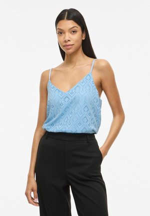 Femme aux longs cheveux foncés portant un haut sans manches bleu clair à motifs et un pantalon noir taille haute sur fond uni.