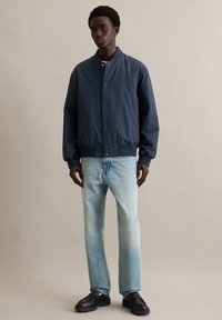 Granatowa kurtka bomberka z zamkiem z przodu, rękawami zakończonymi mankietami i bocznymi kieszeniami, zestawiona z jasnoniebieskimi dżinsami i czarnymi butami.