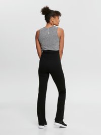 Kvinne med krøllete hår i en knute, iført en grå ermeløs topp, svarte høyhyllede bukser og svarte joggesko, stående med ryggen mot.