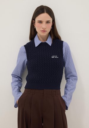 Femme aux longs cheveux bruns porte un pull sans manches en maille torsadée bleu marine sur une chemise rayée bleue et un pantalon plissé marron taille haute.