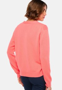 Femme aux cheveux bruns courts portant un pull à manches longues rose corail et un pantalon sombre, de dos avec la tête tournée vers la droite.