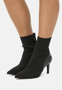 Stivaletti neri alla caviglia con parte superiore in maglia, punta affusolata e tacco a stiletto. Presentano pannelli laterali testurizzati e un dettaglio logo minimalista.