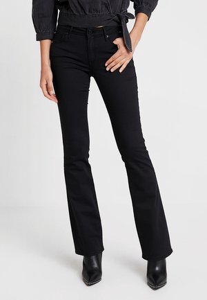 Kobieta ubrana w czarne dzwoniaste dżinsy, czarne botki z noskiem w szpic oraz czarną bluzkę z długim rękawem zawiązaną w talii, stojąca z jedną ręką na biodrze.