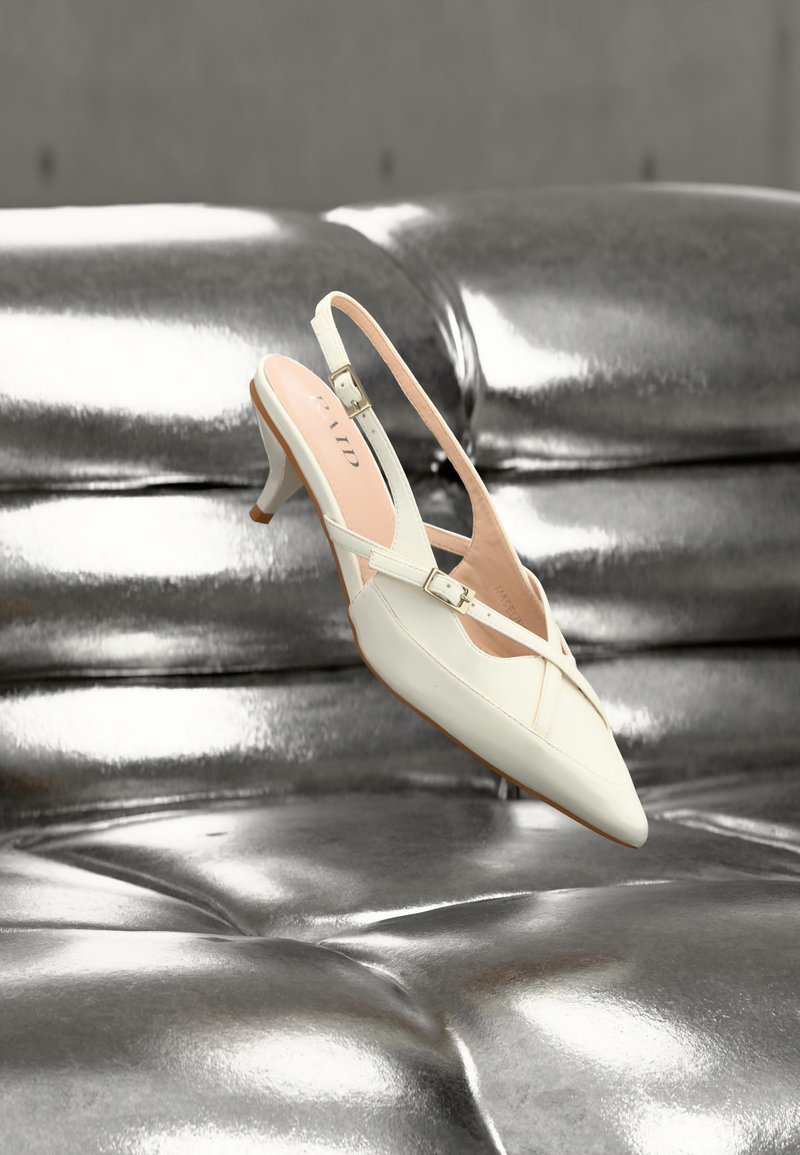 Chaussure blanche à talon kitten slingback avec brides à boucle reposant sur une surface capitonnée en cuir argenté brillant.