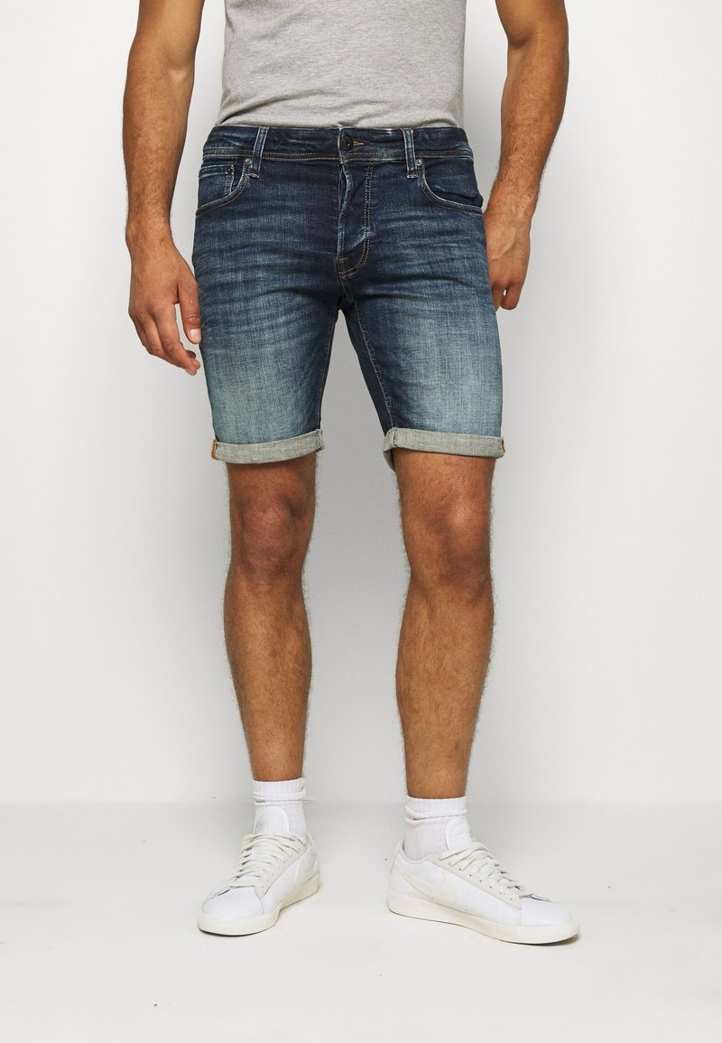 Shorts de denim en azul oscuro con un acabado desgastado y dobladillos rematados. Presenta un estilo clásico de cinco bolsillos y una cremallera con un cierre de botón.