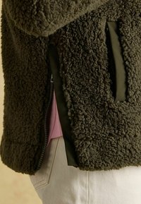 Kurtka polarowa w oliwkowej zieleni z fakturowaną powierzchnią, bocznymi kieszeniami i krótkim zamkiem błyskawicznym. Wnętrze ma kontrastującą różową warstwę pod spodem.