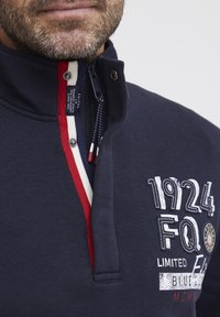 Pullover blu navy con colletto alto, caratterizzato da dettagli a righe rosse e bianche. Include loghi e numeri ricamati, chiusura con zip e hardware decorativo.