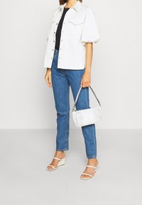 Camisa blanca de gran tamaño con mangas abullonadas, top negro, jeans de pierna recta azules y un bolso blanco texturizado. Sandalias con tacones de tiras beige.