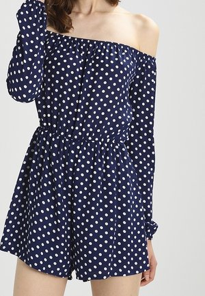 Vrouw die een marineblauwe off-shoulder romper draagt met witte polkadots, lange mouwen en een elastische taille, staand tegen een effen achtergrond.