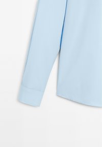 Camicia a maniche lunghe azzurro chiaro con texture liscia, polsini standard e orlo arcuato. Il tessuto appare leggero e arioso.