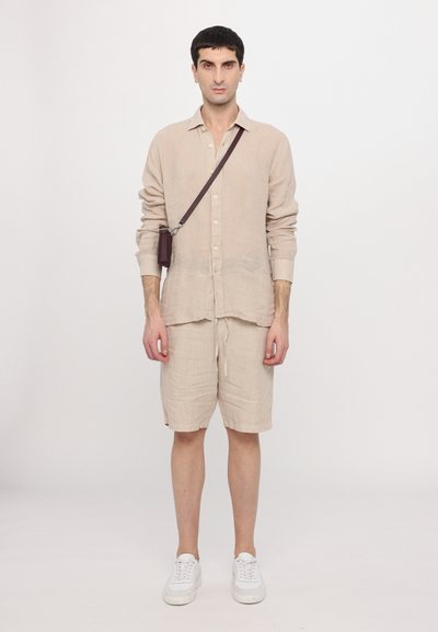 Camisa de linho bege com botões e mangas longas, combinada com calções bege. Ténis brancos e uma mala de ombro castanha completam o conjunto.