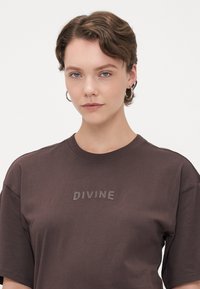 Hnědá bavlněná tričko s kulatým výstřihem a krátkými rukávy, s texturovaným logem "DIVINE" na přední straně. Hladký povrch látky.