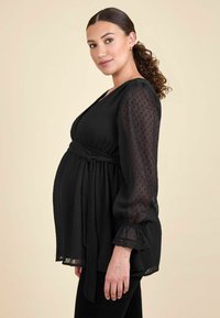 Čierna tehotenská blúzka s prekrývajúcim dizajnom, textúrovaná tkanina, dlhé priesvitné rukávy a volánikové manžety. Obsahuje viazanie v páse.