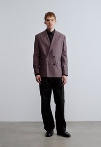 Doppelreihiges mauvefarbenes Sakko mit scharfen Revers, kombiniert mit einem schwarzen Hemd und weiten schwarzen Hosen, abgerundet mit schwarzen Schuhen. Glatte Textur.