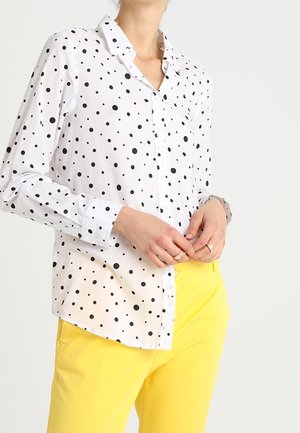 Woman wearing a white long-sleeve shirt with black polka dots and bright yellow pants, hands clasped near waist.