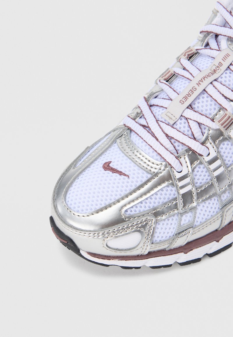 Chaussure de sport argentée et blanche en matériau mesh, avec un système de laçage complexe et un logo Nike swoosh notable sur le côté. Semelle en caoutchouc.