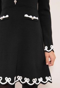 Rochie neagră tricotată cu accente albe cu model valurat și tiv cu bucle la mâneci și poale, cu nasturi decorativi pe buzunarele de la piept.