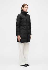 Γυναίκα που φοράει μαύρο παλτό με καπιτονέ σχεδιασμό και ψηλό γιακά, λευκό παντελόνι με φαρδύ πόδι, μαύρα μυτερά παπούτσια, στέκεται μπροστά σε απλό λευκό φόντο.