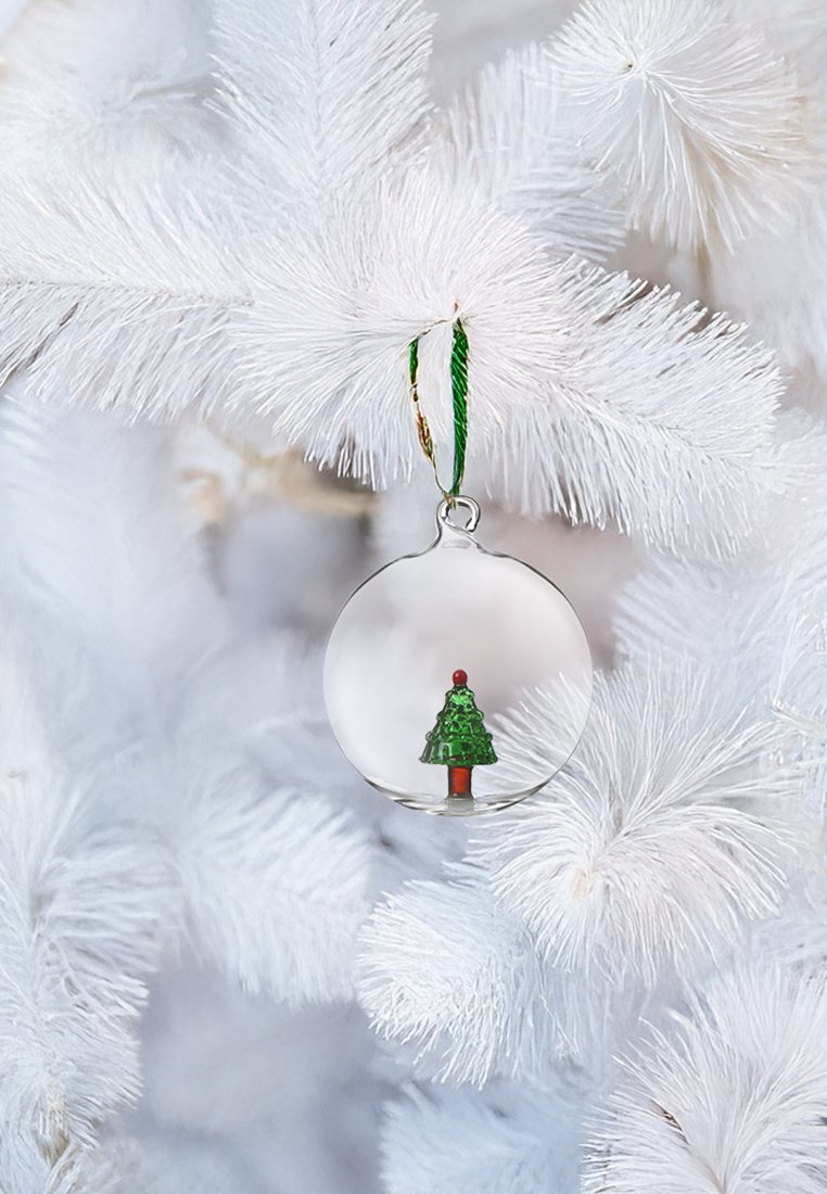 Przezroczysta szklana ozdoba z miniaturową zieloną choinką i czerwonymi detalami, zawieszona na zielonej wstążce na białej sztucznej choince.
