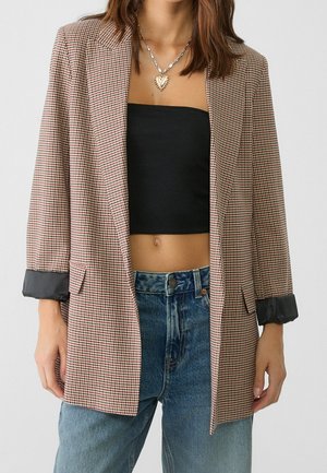 Kvinde iført en ternet blazer med sorte satinmanchetter, sort stropløs crop top, blå jeans og en guldhalskæde med hjertevedhæng.