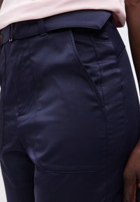 Κοντινό πλάνο σε καλοραμμένο παντελόνι σκούρου μπλε με πλαϊνές τσέπες και θηλιές για ζώνη, φορεμένο με ροζ ανοιχτό πουκάμισο, που δείχνει μέρος του χεριού ενός ατόμου.