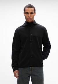 Camisa cuspida preta com fecho de botões, detalhe de bolsa no peito e bolsos laterais. Apresenta uma textura suave e um corte descontraído.