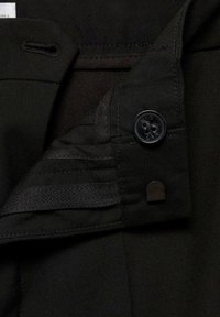 Crni materijal s vidljivim šavovima, dugme s logotipom i crna tekstilna petlja. Sadrži suptilne teksture i slojevite dizajnerske detalje.
