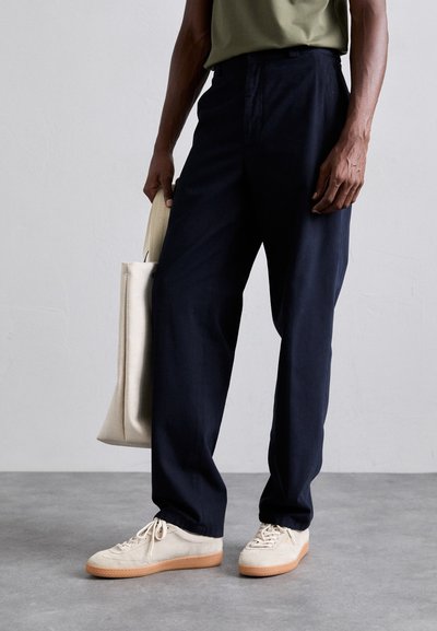 Marinblå vida byxor med en slät textur, parat med krämfärgade snörskor och en beige tote-väska. Minimalistisk design och rena linjer.