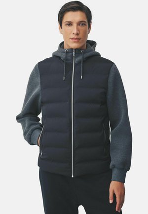 Joven vestido con un chaleco acolchado negro sobre una sudadera con capucha gris oscuro, con las manos en los bolsillos del chaleco, mirando de frente sobre un fondo blanco.