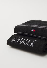 Calcetines negros con el texto en blanco "TOMMY HILFIGER". Presentan rayas grises y un pequeño logo en rojo, blanco y azul en la esquina. Textura de punto suave.
