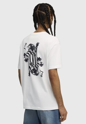 Person with braided hair wearing a white T-shirt featuring a black and white graphic of mirrored roaring panthers and soccer balls on the back.