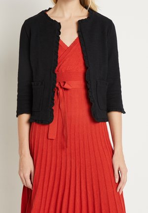 Schwarze verkürzte Strickjacke mit Rüschensaum, ausgestattet mit zwei vorderen Taschen, kombiniert mit einem roten, plissierten Wickelkleid. Weicher Stoff, lässiges Design.