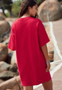 Rød kortærmet kjole med lige snit, lavet af blødt stof. Har en afslappet pasform og enkelt design, vist bagfra mod en baggrund af klipper.