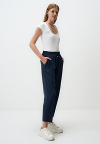 Witte aansluitende bodysuit, marineblauwe broek met elastische taille en zakken, taps toelopende pijpen en klassieke witte sneakers met textuurzolen en kleine accenten.