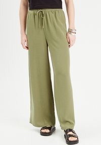 Pantalons taille large vert olive en matériau doux, dotés d'une taille élastique avec cordon de serrage. Associés à des sandales noires.