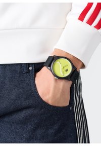 Must musta kell, millel on ümmargune roheline nägu, hõbedased käed ja logo, kantud randmel. Taustal on valge spordipusa ja tummad teksased.