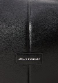 Melna ādas somiņa ar teksturētu virsmu, vertikālām šuvēm un etiķeti, kurā rakstīts "ARMANI EXCHANGE" baltā krāsā.