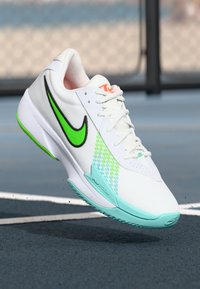 Hvide Nike løbesko med grøn swoosh, teal sål og blå-grøn mønstret side, svævende over en tennisbane.
