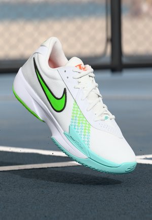 Bijela Nike tenisica za trčanje s zelenim swoosh-om, tirkiznom potplatom i bočnoj strani s plavo-zelenim uzorkom, lebdi iznad površine teniskog terena.