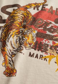 Tissu de couleur claire présentant un motif de tigre orné de strass multicolores. Des accents rouges et marron notables complètent le motif.