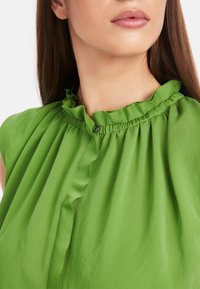 Grüne Bluse mit rüschendem Ausschnitt und einem Knopfdetail. Der Stoff wirkt weich mit subtilen Drapierungen, die die Gesamtstruktur unterstreichen.