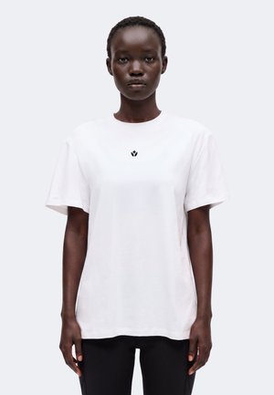 Femme aux cheveux courts portant un t-shirt blanc uni avec un petit logo noir au centre, debout devant un fond gris clair.