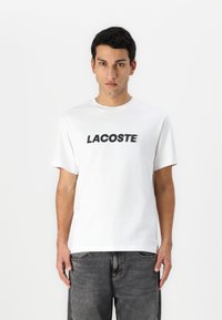 Baltos medvilnės marškinėliai su ryškiu juodu "LACOSTE" logotipu priekyje. Trumpomis rankovėmis ir apvaliu iškirpimu. Minimalistinis dizainas.