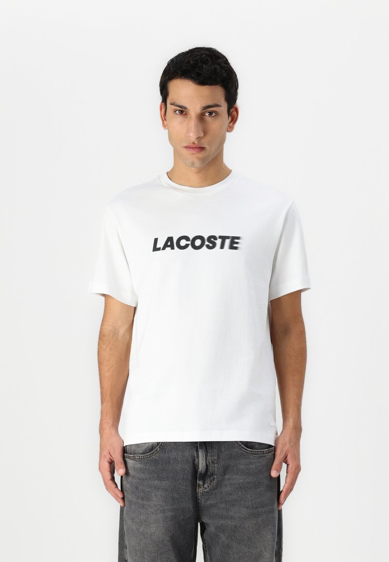 Baltos medvilnės marškinėliai su ryškiu juodu "LACOSTE" logotipu priekyje. Trumpomis rankovėmis ir apvaliu iškirpimu. Minimalistinis dizainas.