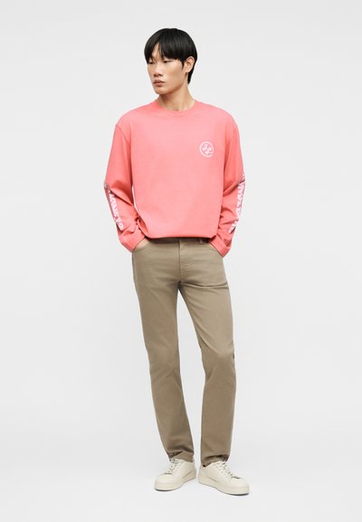 Νέος άνδρας που φοράει κοραλί μπλούζα με μακριά μανίκια και λευκά γράμματα στα μανίκια, μπεζ παντελόνι και λευκά αθλητικά παπούτσια, στέκεται με τα χέρια στις τσέπες.